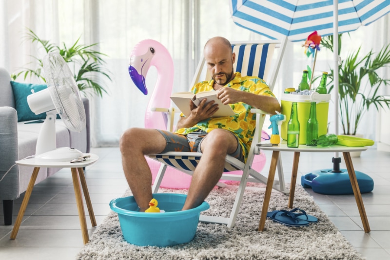 Man sitting in a beach scene in his home. Sand, beach chair, umbrella, kitty pool.