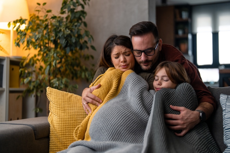 Family cuddled up under blankets on couch wondering why their furnace isn't heating their home.